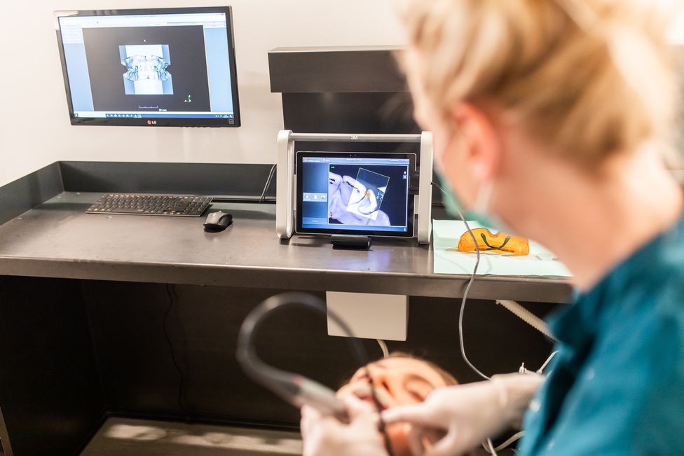Oral hygienist works in a patient's mouth. She looks up at a 3D image of the teeth on the screen in front of her.